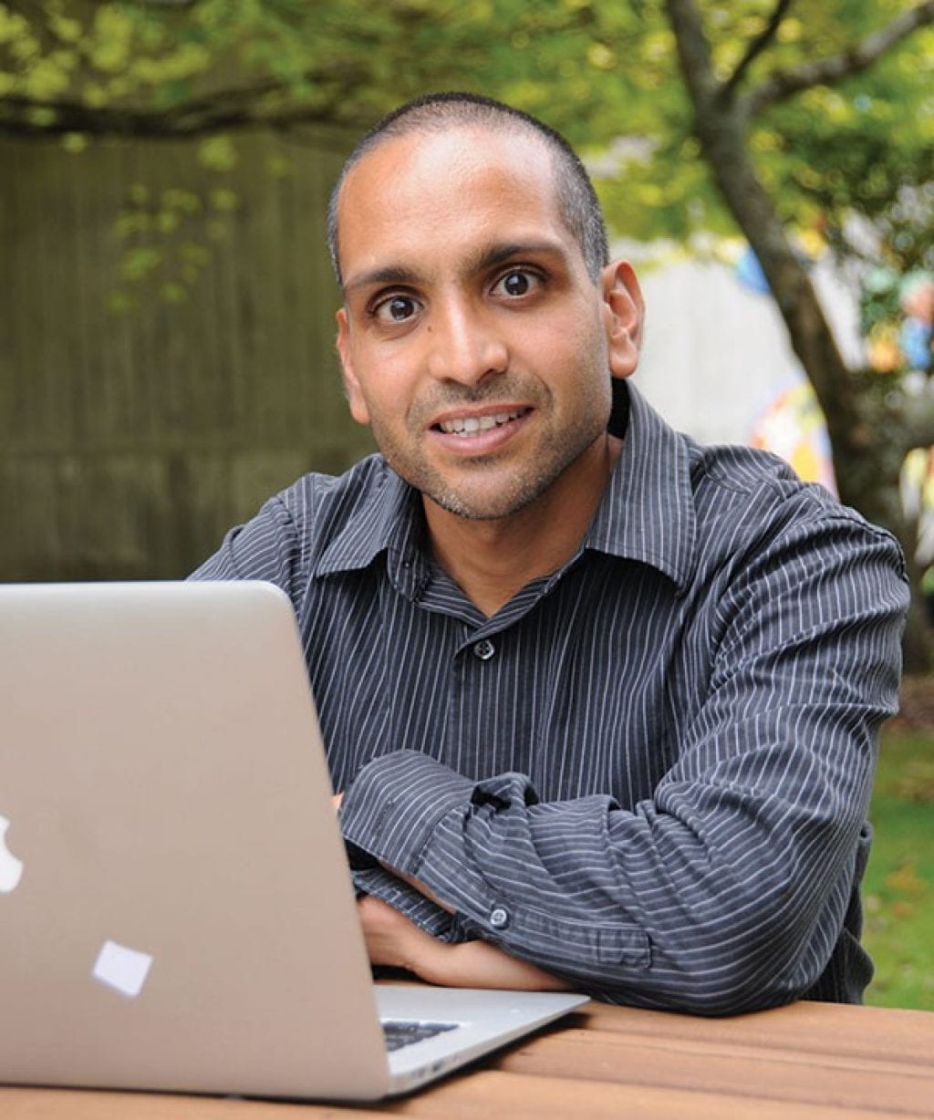 Dr. Shah sits outside on a table in front of a laptop looking at the camera